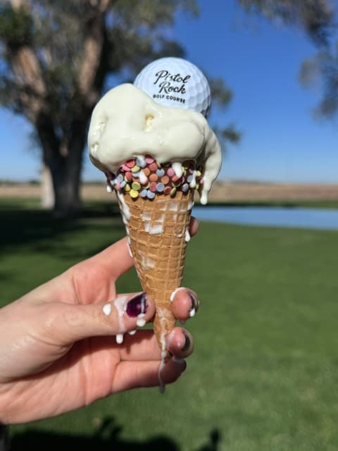 A dipped waffle cone rolled in candy pieces and topped with a Pistol Rock golf ball, held up beside the course
