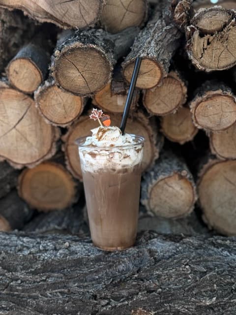 An iced fall drink topped with whipped cream and a maple leaf pick, in front of a stack of firewood